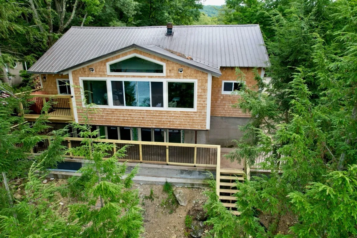 The exterior of the lodge is showcased, with large front windows and a wooden deck surrounded by lush greenery. The inviting deck is ideal for outdoor relaxation, offering views of the natural landscape and an immediate connection with the lakefront environment.