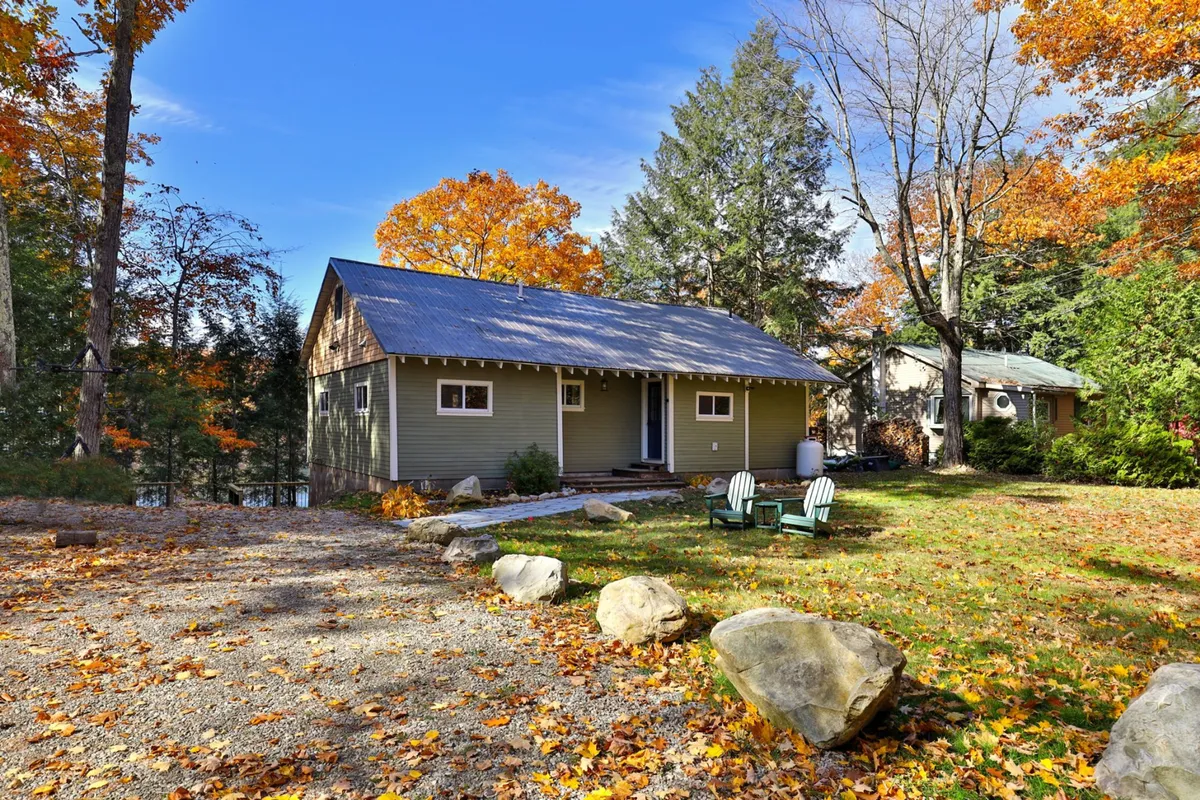This exterior image shows the house surrounded by vibrant autumn trees, Adirondack chairs arranged on large rocks, and a cozy outdoor setting perfect for gathering. The scene captures the warmth of outdoor relaxation and the inviting atmosphere of a small-town getaway.