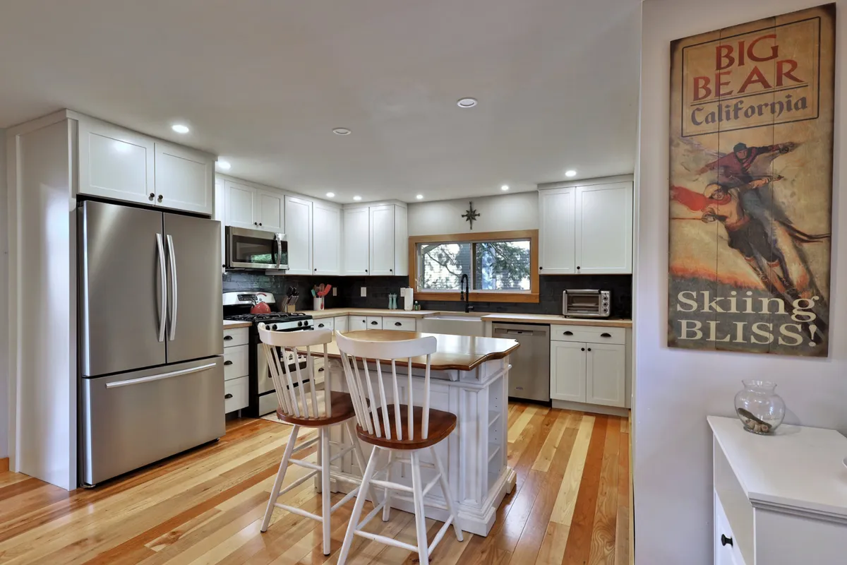 A bright, welcoming kitchen featuring a spacious island, gleaming stainless steel appliances, a window offering a scenic view, and charming details like a vintage skiing poster. The setup is ideal for gathering while prepping meals or enjoying casual conversation.