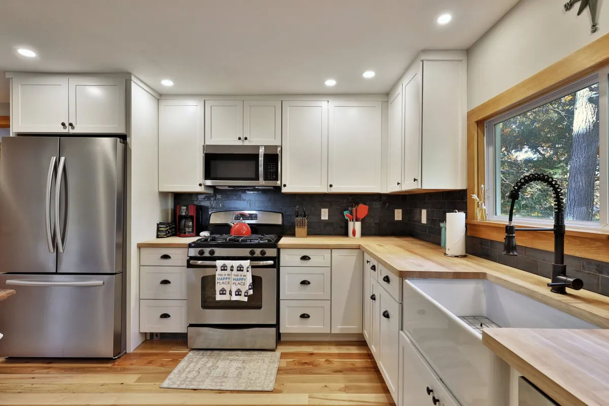 A cozy kitchen corner with a prominent stainless steel refrigerator and gas stove, complemented by a large window bathing wooden countertops in natural light. The inviting setup suggests both functionality and warmth, ideal for cooking and gathering.