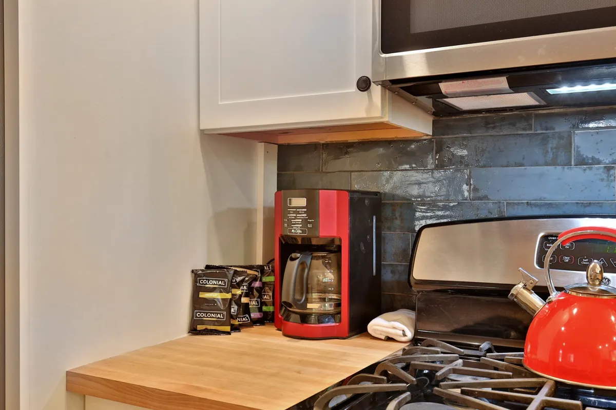 This image features a functional kitchen corner with a coffee maker, red kettle, sleek countertops, and a stylish backsplash. The area is well-lit and organized, offering space for meal prep and casual gatherings, and hinting at modern, user-friendly appliances.
