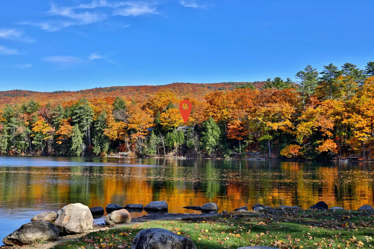 A tranquil scene featuring a calm lake surrounded by vibrant autumn foliage, large boulders along the shore, and distant hills under a clear sky. The reflective water surface enhances the beauty of the changing leaves, making it an ideal spot for outdoor recreation such as hiking, fishing, and photography.