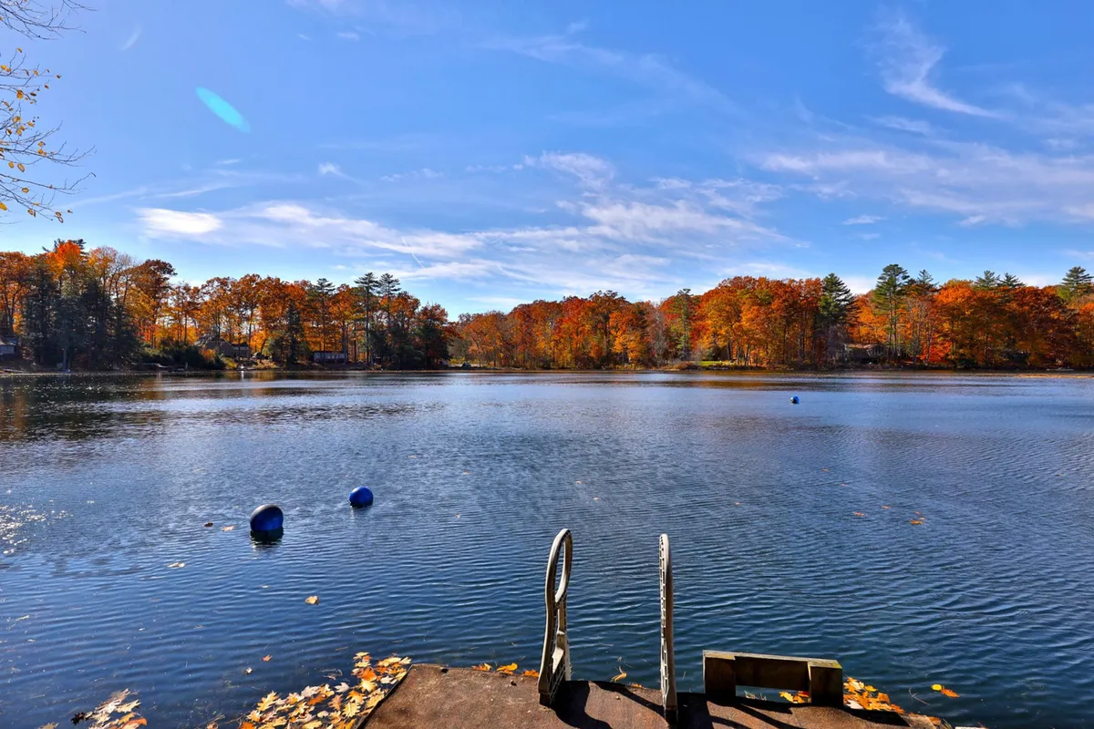 A serene lake scene with a wooden dock extending over the water, vibrant autumn trees along the shore, and blue buoys bobbing in the gentle ripples. The tranquil setting invites guests to swim, boat, fish, or simply relax by the water.