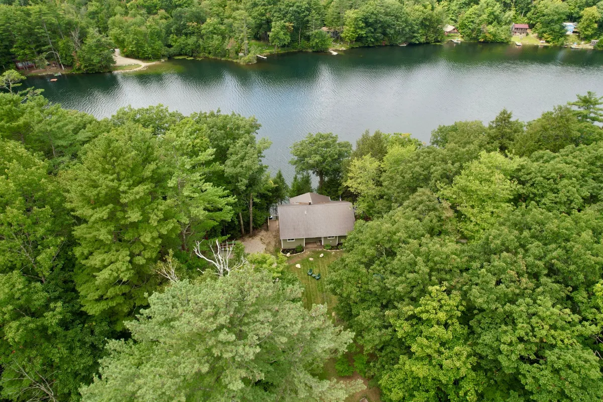The image showcases a charming lakefront house nestled among dense trees, with inviting outdoor seating overlooking the tranquil waters of Fern Lake. The scene evokes relaxation and connection to nature, with the lake, the house, and the lush Vermont landscape all visible.