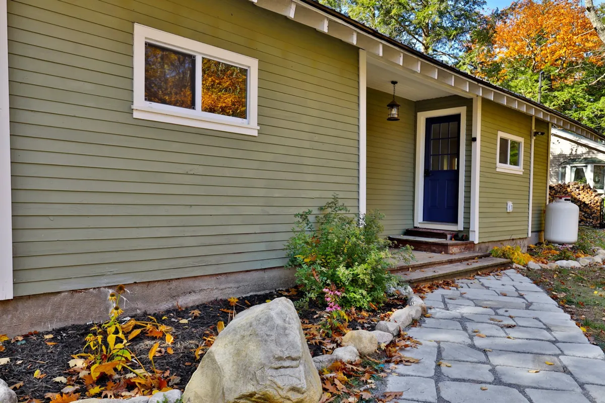 Exterior shot featuring the house with green siding, a blue front door, pathway stones, garden plants, and a large rock. This entryway scene conveys a sense of arrival and sets the stage for adventures into the surrounding small towns and cultural attractions.