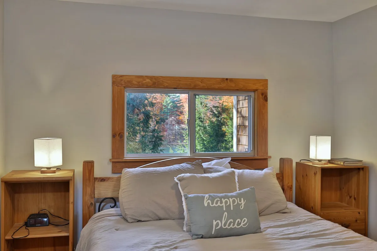 A welcoming bedroom with a bed dressed in decorative pillows, a large window offering tranquil views, and matching nightstands with lamps. The ambiance is peaceful and inviting, suggesting restful sleep and morning relaxation.