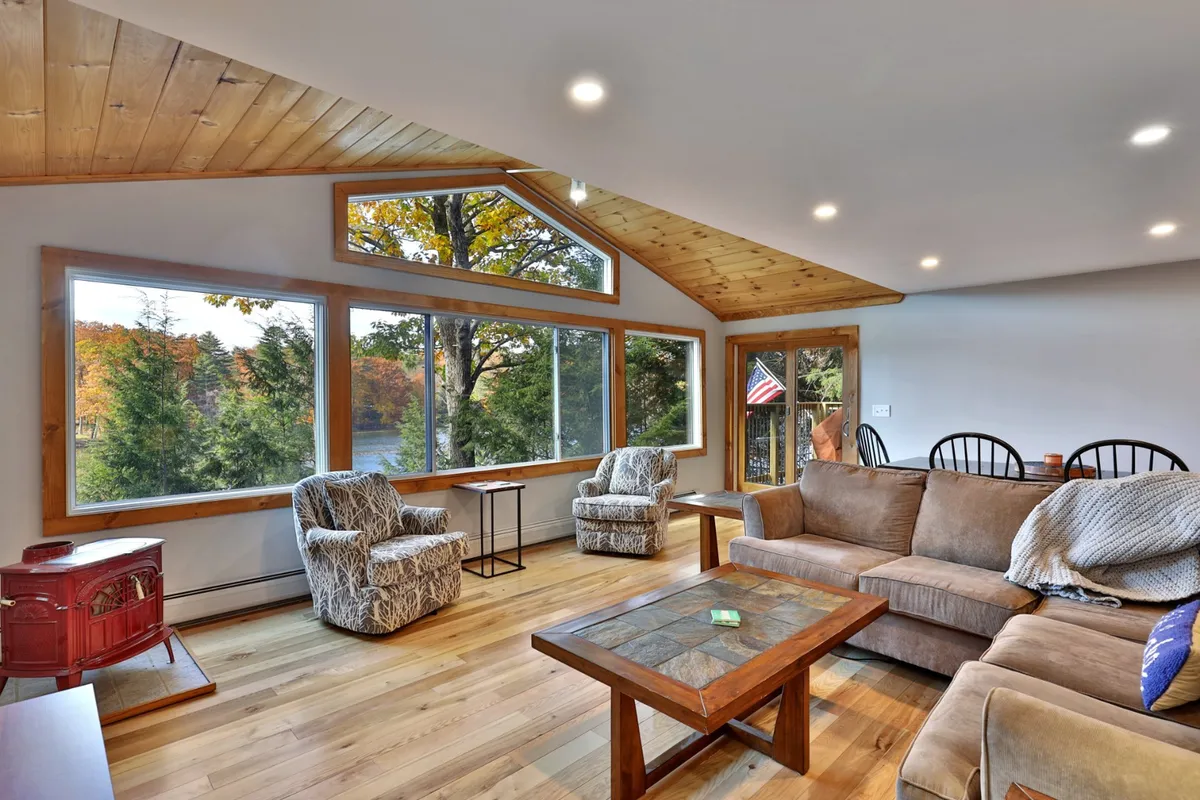 A warm, rustic living space with a wooden ceiling, large windows revealing scenic lake views, comfortable seating, a wooden coffee table, and a charming red stove. The inviting atmosphere is ideal for movie nights, family games, or relaxing with the picturesque outdoors always in sight.